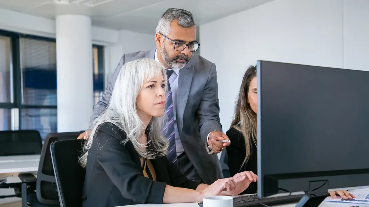 A team lead pointing at the screen while an assistant works through a task at her desk