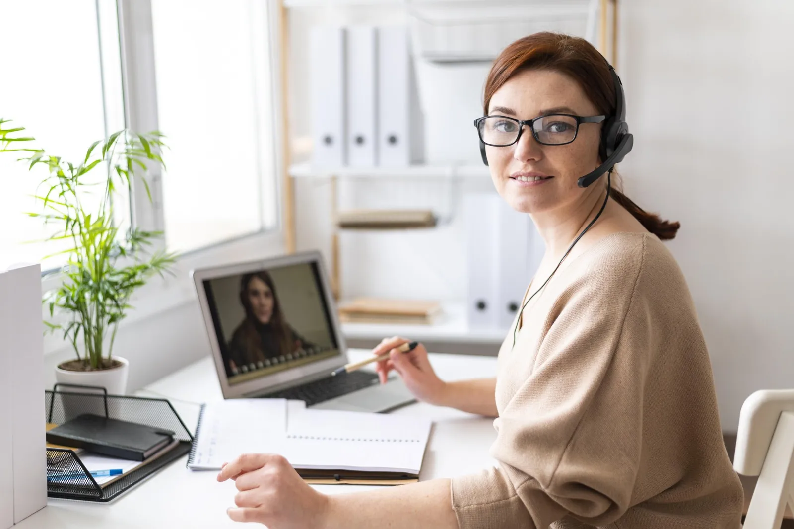 An assistant wearing a headset on a video call, taking notes at her desk during onboarding
