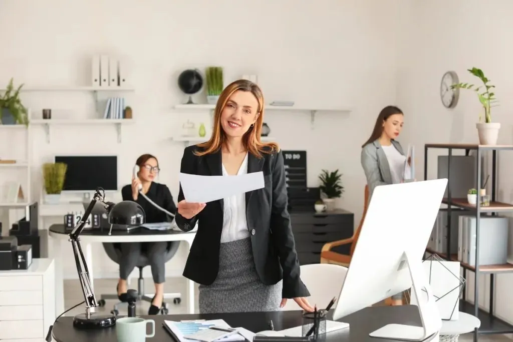 A smiling businesswoman holding a document in a busy office, colleagues on the phone behind her