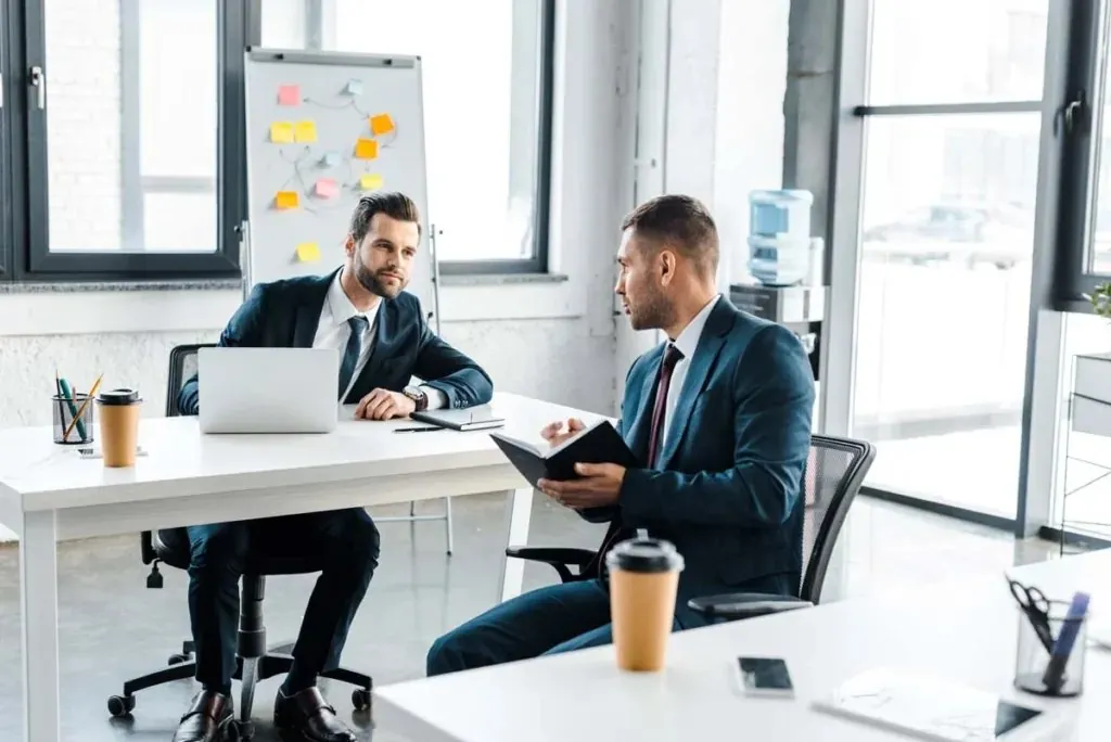 Two businessmen in suits talking across desks in a bright office